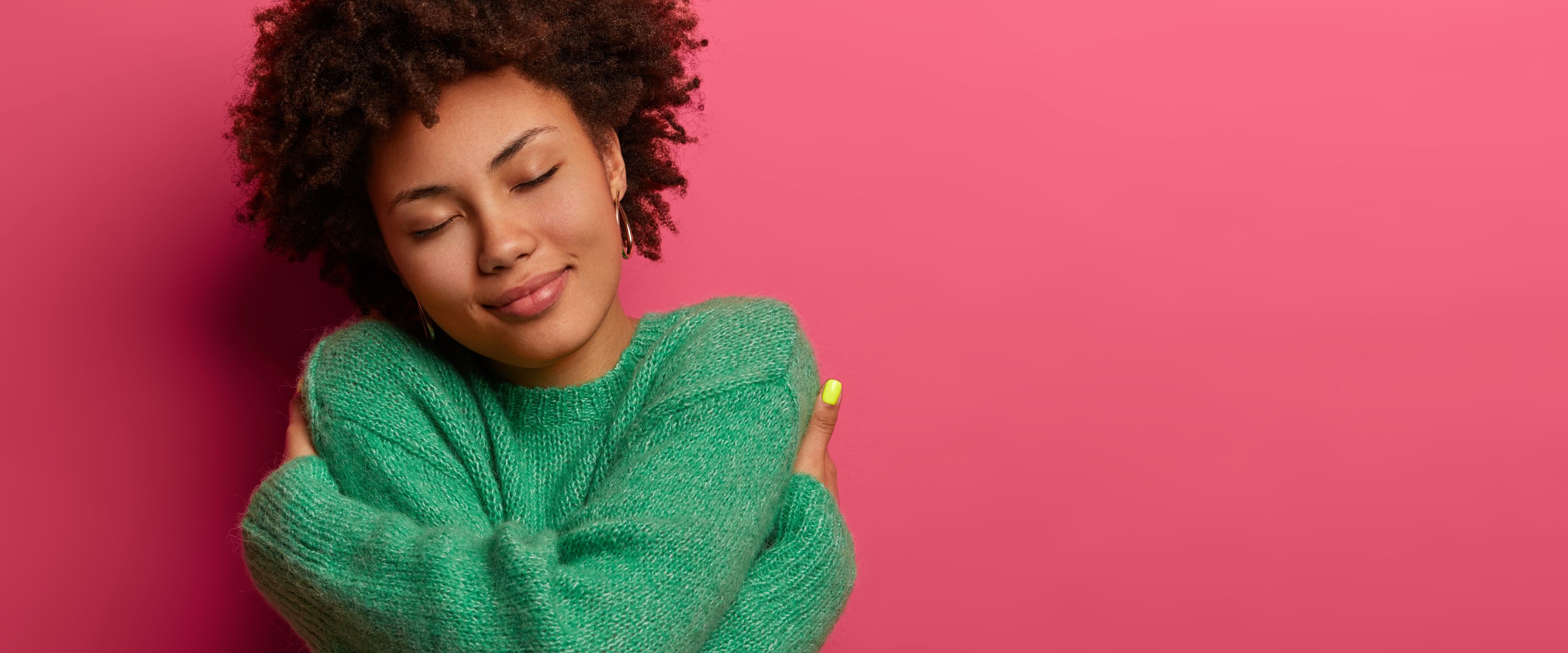Women in a green sweater hugging herself in front of a pink background