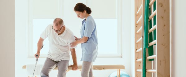 Female physiotherapist assisting elderly man with mobility training in a bright rehabilitation clinic. Ideal for senior physical therapy, stroke recovery, and in-home care services.
