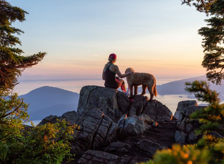 Woman and her dog sitting on a large rock over looking mountains at sunset