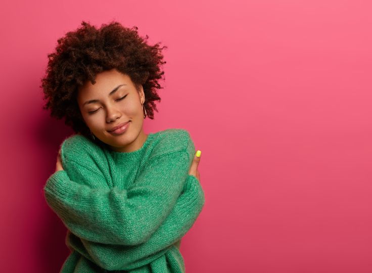 Women in a green sweater hugging herself in front of a pink background